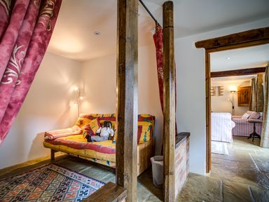 A small sitting area with a futon sofa bed and a doll on it next to wooden beams and red curtains at Little Barn in Ascott near Chipping Norton