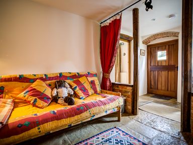A sitting area with a colorful sofa and stuffed toy near a rustic wooden door with a red curtain at Little Barn in Ascott near Chipping Norton