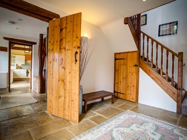 A hallway with wooden doors a wooden stair railing a bench and a kitchen visible through an open doorway at Little Barn in Ascott near Chipping Norton