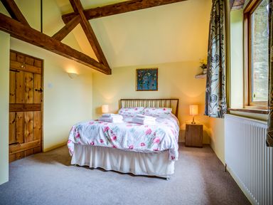 A bedroom with a wooden bed and floral bedding two bedside tables with lamps a wooden door and window with patterned curtains at Little Barn in Ascott near Chipping Norton