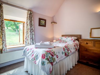 A small bedroom with a single bed with floral bedding next to a window with floral curtains and a wooden dresser at Little Barn in Ascott near Chipping Norton