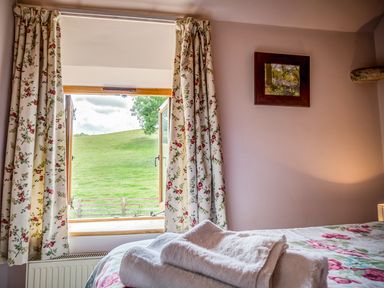 A bedroom with floral curtains and bedding a window opening to a green field and towels on a bed at Little Barn in Ascott near Chipping Norton
