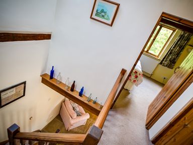 A hallway with a staircase, bottles on a wooden shelf, and a glimpse of a bedroom with a bed and window at Little Barn in Ascott near Chipping Norton
