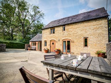 A stone cottage with wooden doors and windows and outdoor table with mugs at Little Barn in Ascott near Chipping Norton
