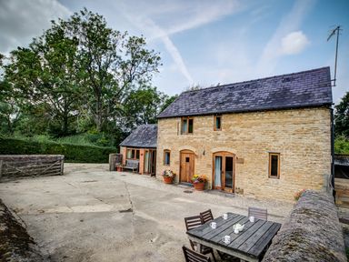 An outdoor patio area with a wooden table and chairs with mugs on the table next to a stone house at Little Barn in Ascott near Chipping Norton