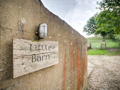 A wooden sign reading Little Barn mounted on a concrete wall next to a gravel driveway with a field and trees in the background at Little Barn Ascott near Chipping Norton