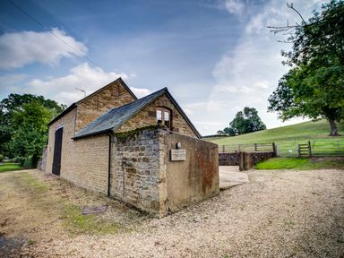 A stone barn with a slate roof next to a gravel driveway and green fields at Little Barn in Ascott near Chipping Norton