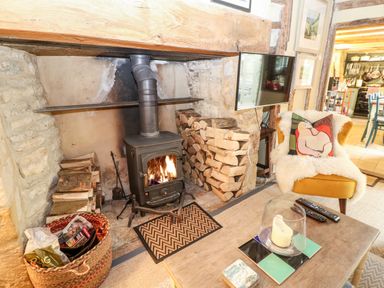 A wood burning stove with fire logs beside it a basket a chair with cushions a wooden table with a candle and remotes at Benfield in Stow-on-the-Wold