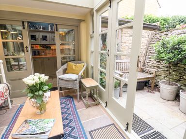 A sunroom with a wicker chair yellow pillow small table wooden coffee table with flowers magazine and a view of a stone patio with a wooden bench at Benfield in Stow-on-the-Wold