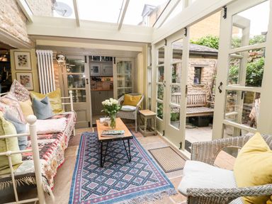A sunroom with a sofa with cushions a coffee table with flowers wicker chairs and glass doors leading to a patio at Benfield in Stow-on-the-Wold