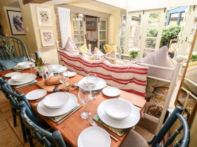 A dining table set with plates glasses and bread next to a sitting area with cushions and glass doors in a room at Benfield in Stow-on-the-Wold