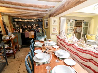 A kitchen and dining area with a wooden table set with plates glasses and bread and a seating area with a striped sofa and cushions at Benfield in Stow-on-the-Wold