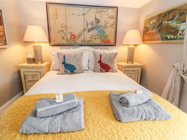 A bedroom with a bed with white sheets and yellow blanket two bedside tables with lamps and quilts with rabbit designs on cushions at Benfield in Stow-on-the-Wold