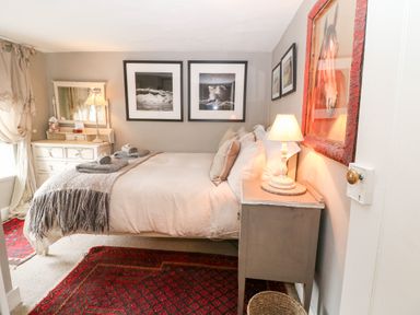 A bedroom with a bed, bedside table with a lamp, dresser with a mirror, framed pictures on walls, and red patterned rugs at Benfield in Stow-on-the-Wold