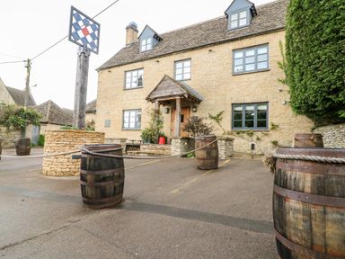 A stone building with multiple windows wooden barrels with ropes and a signpost outside at Anvil Cottage in Chipping Norton