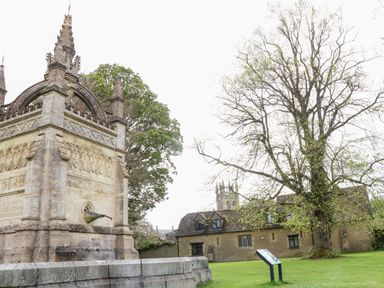 An outdoor scene with a stone monument with water spout and historic buildings with trees at Anvil Cottage in Chipping Norton