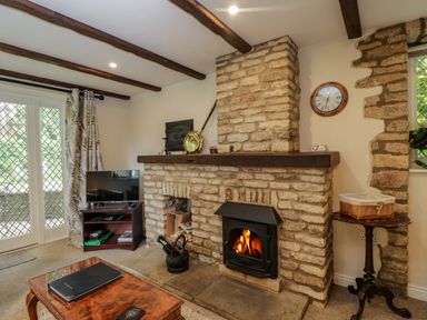 A living room with a stone fireplace wooden beams a television on a stand and a coffee table at Anvil Cottage in Chipping Norton