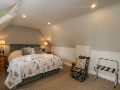 A bedroom with a double bed, two bedside tables with lamps, a floral armchair with cushions, and a luggage rack at Anvil Cottage in Chipping Norton