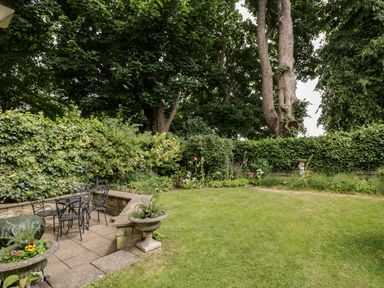 A garden with metal chairs and table on a paved area and plants around a lawn at Anvil Cottage in Chipping Norton