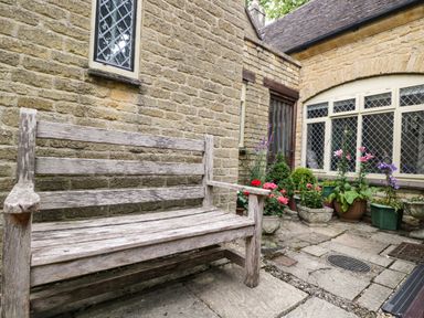 An outdoor stone patio with a wooden bench and various potted plants next to a stone building with diamond-patterned windows at Anvil Cottage in Chipping Norton