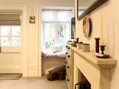 A room with a window seat with cushions a door a chest of drawers a fireplace with candles and plants and a stuffed hippopotamus at Hare House in Chipping Norton