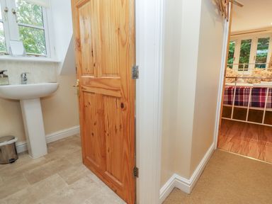 A bathroom with a sink and wooden door next to a hallway leading to a bedroom with a metal bedframe and plaid bedding at The Cottage At Barrow Mead in Bath