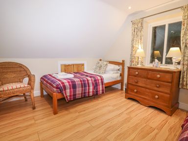 A bedroom with a single bed covered with a plaid blanket a wooden chest of drawers with two lamps and a wicker chair at The Cottage At Barrow Mead in Bath