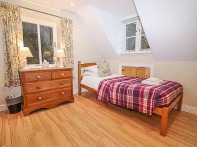 A bedroom with a single bed covered with a plaid blanket wooden dresser with two lamps and floral curtains at The Cottage At Barrow Mead in Bath