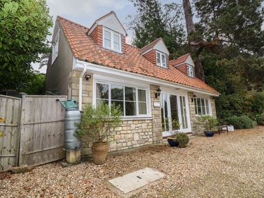 A stone cottage with a red tiled roof and three dormer windows surrounded by plants and gravel at The Cottage At Barrow Mead in Bath