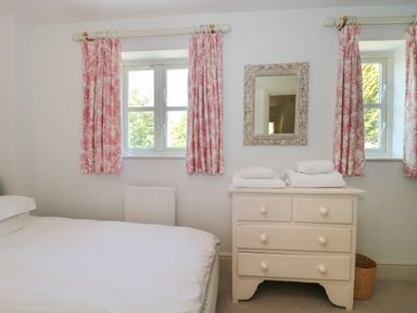 A bedroom with bed white chest of drawers mirror and windows with pink curtains at Batemans in Nailsworth