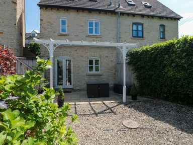 A stone house with white framed windows and doors a white pergola and a gravel and paved garden with plants at Batemans Nailsworth