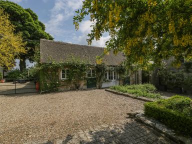 A stone building with climbing plants and a gravel courtyard with trees and shrubs at Hazel Manor Wing in Painswick