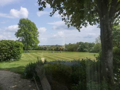 A view of a lawn with trees and shrubs under a partly cloudy sky at Hazel Manor Wing in Painswick