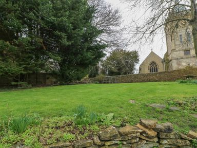 A garden with grass and stone wall with trees and a church building in the background at Newbury Cottage in Bourton-On-The-Water