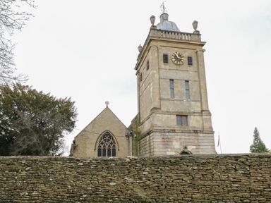 A stone church with a clock tower behind a stone wall with trees on the left at Newbury Cottage in Bourton-On-The-Water