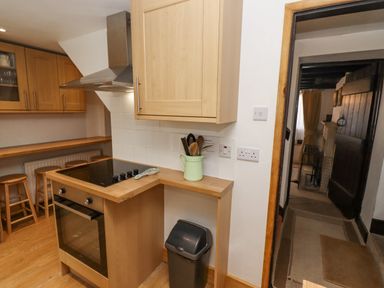 A kitchen with wooden cabinets a black stove a trash bin and a doorway leading to another room at Newbury Cottage in Bourton-On-The-Water