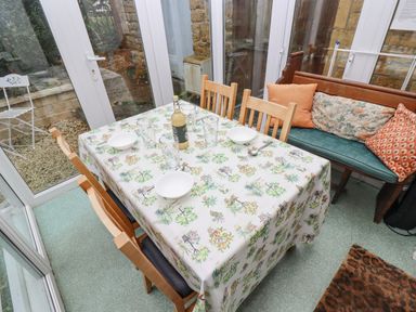 A dining table with a floral tablecloth set with bowls glasses and a bottle in a room with wooden chairs and a bench with cushions at Newbury Cottage in Bourton-On-The-Water