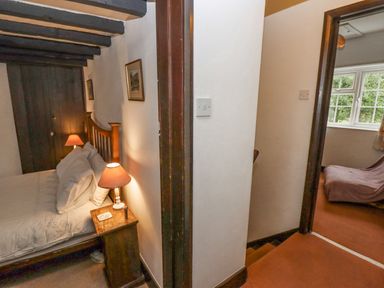 A bedroom with a bed lamp and bedside table next to a hallway leading to another room with a window and a covered chair at Newbury Cottage in Bourton-On-The-Water