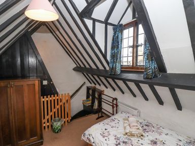 A bedroom with a bed, wooden wardrobe, window with blue curtains, and exposed black ceiling beams at Newbury Cottage in Bourton-On-The-Water