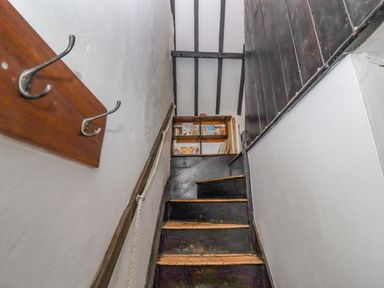 A narrow staircase with black wooden steps and white walls with a wooden coat rack and shelf with books at Newbury Cottage in Bourton-On-The-Water