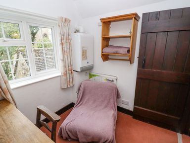 A small room with a wooden chair and desk a window with floral curtains a radiator covered with a blanket a wall-mounted shelf with a folded blanket and a dark wooden door at Newbury Cottage in Bourton-On-The-Water