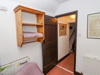 A small room with a wooden wall shelf holding a towel a dark wooden door open to a hallway with a wall hanging and a radiator on the right at Newbury Cottage in Bourton-On-The-Water