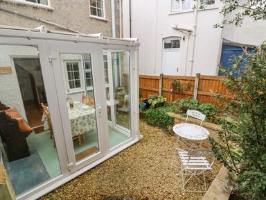 An outdoor patio area with a white metal table and two chairs next to a wooden fence and a glass conservatory with a dining table inside at Newbury Cottage in Bourton-On-The-Water