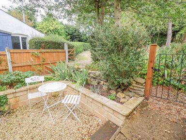 A small garden area with a white metal table and two chairs on gravel next to a stone planter with plants and a wooden fence with a metal gate at Newbury Cottage in Bourton-On-The-Water