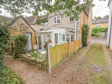 A stone cottage with a small garden and white conservatory next to a gravel driveway at Newbury Cottage in Bourton-On-The-Water