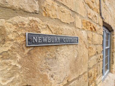 A close up of a stone wall with a metal sign reading Newbury Cottage near a window at Newbury Cottage Bourton-On-The-Water