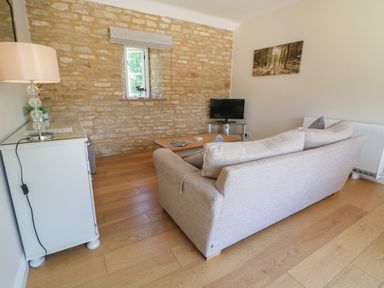 A living room with a beige sofa, wooden coffee table, small TV on a glass stand, white cabinet with a lamp, and stone wall at Saddlebacks Barn in Burford