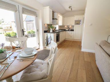 A kitchen with white cabinets and wooden floor next to a dining table set and glass doors at Saddlebacks Barn in Burford
