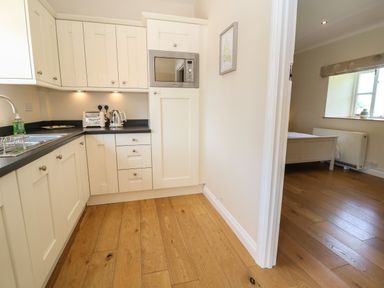 A kitchen with white cabinets a microwave toaster kettle and black countertop with a view into a bedroom with a window at Saddlebacks Barn in Burford