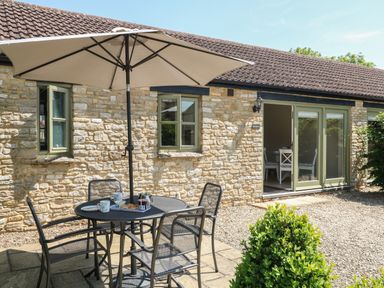 An outdoor patio area with a round table four chairs and a large umbrella outside a stone building with green windows and sliding doors at Saddlebacks Barn in Burford